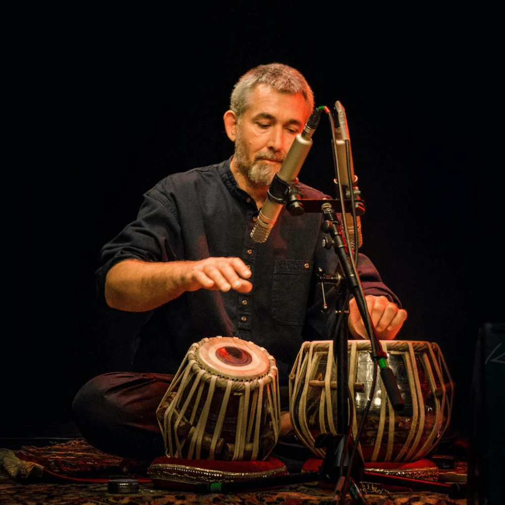 A white man dressed in black sitting down and playing tabla drums